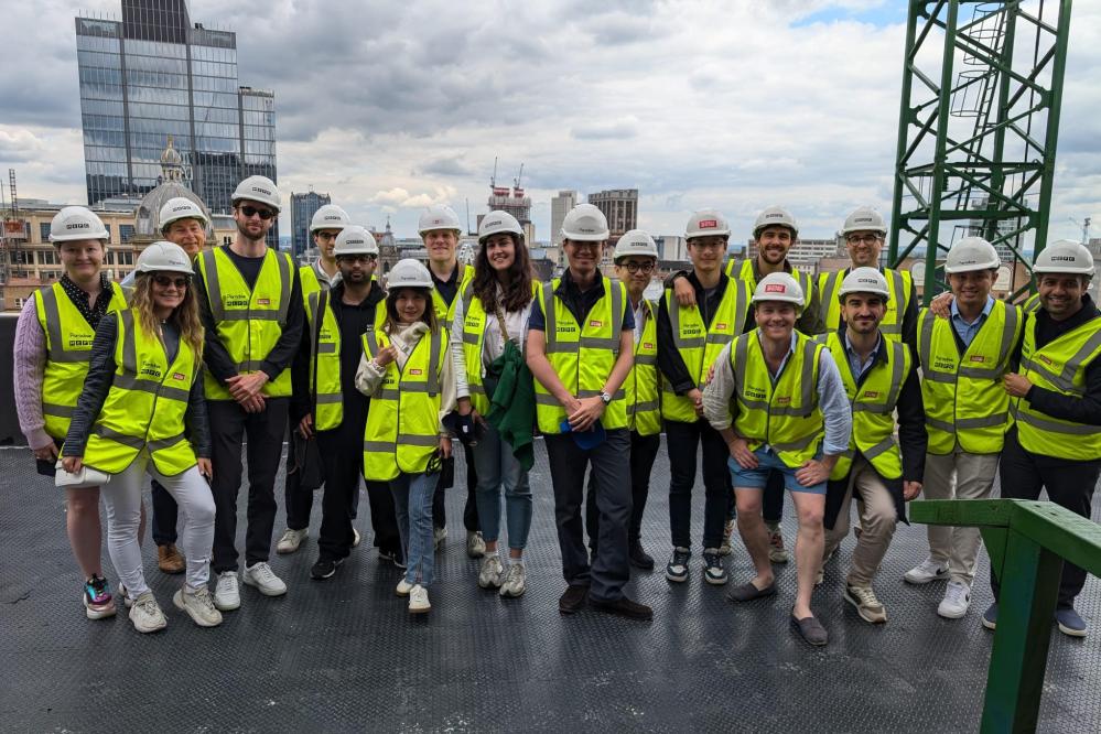 A photograph of the 2023-25 cohort of MSt in Real Estate students visiting 3 Chamberlain Square in Birmingham, UK. The students are wearing yellow high-vis jackets and white hard hats.