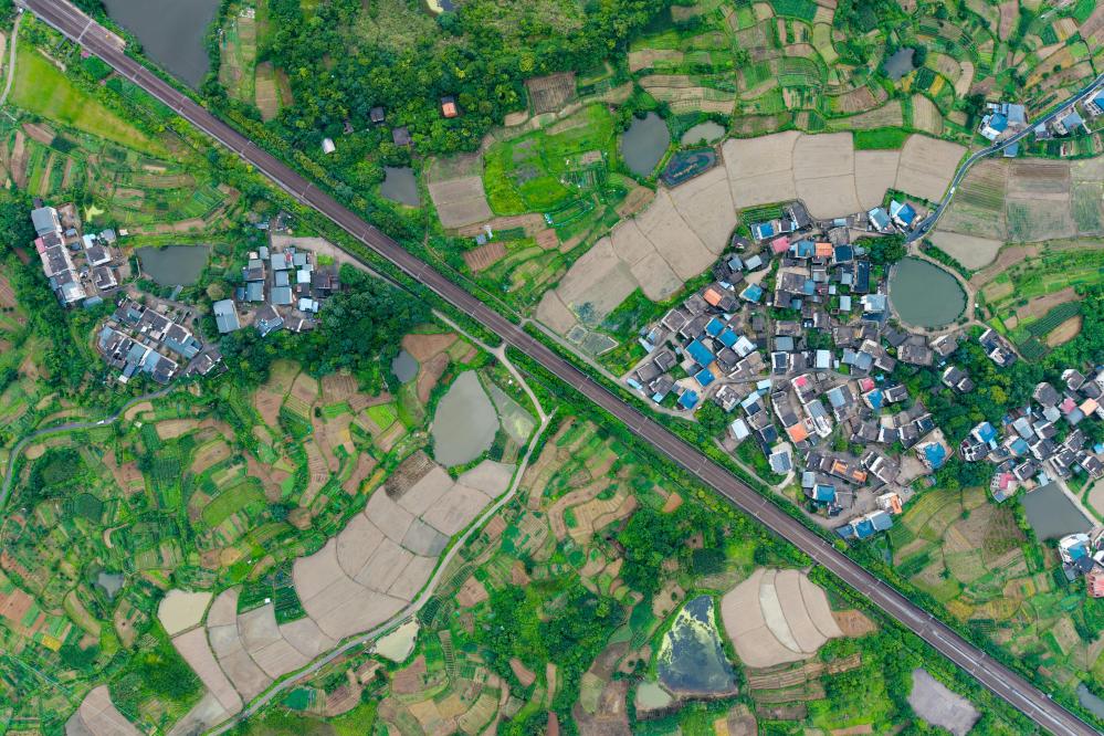 A top-down aerial photograph of a road cutting through green fields, bordered by small buildings